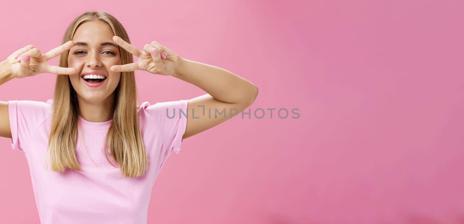 Joyful friendly and peaceful common european woman with blond hair in casual t-shirt showing peace or disco signs over eyes smiling cheerfully at camera having fun against pink background. Copy space