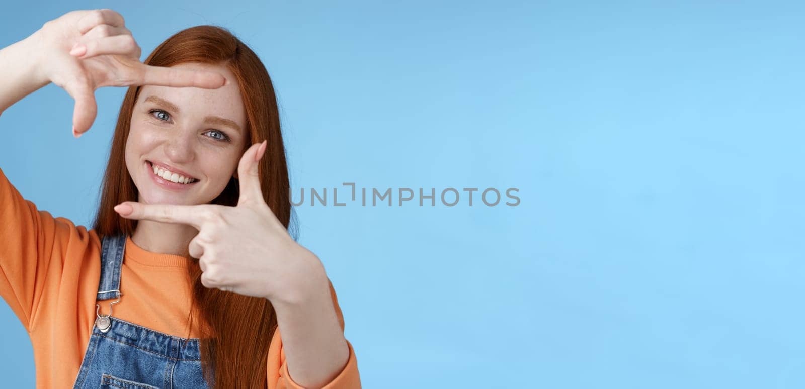 Joyful attractive sincere redhead young girl searching inspiration find perfect angle take good shot make hand frames look through delighted amused smiling broadly white teeth, blue background by Benzoix