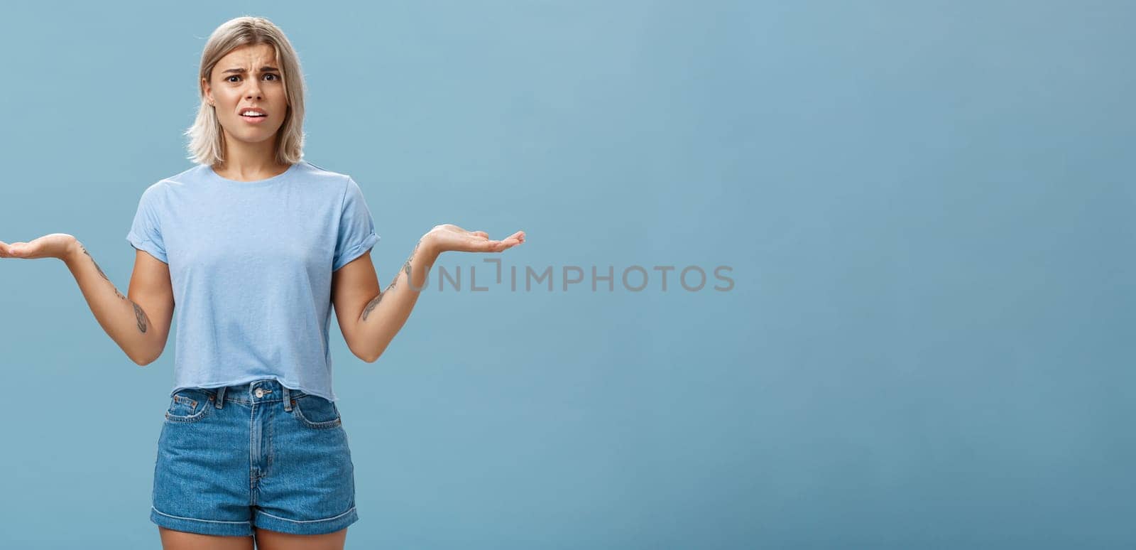 Girl feeling struggles to understand what happening. Confused perplexed intense good-looking female with fair hair shrugging with hands spread aside in displeased clueless pose posing over blue wall.