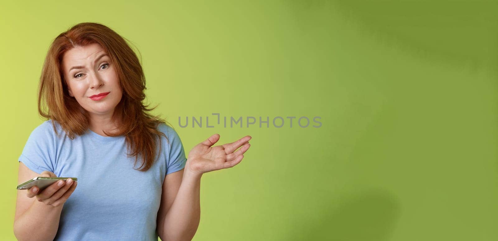 Well meh. Indifferent careless hesitant redhead middle-aged woman mature red female shrugging hold smartphone smirk bored uninterested hold hand aside apathetic attitude green background.