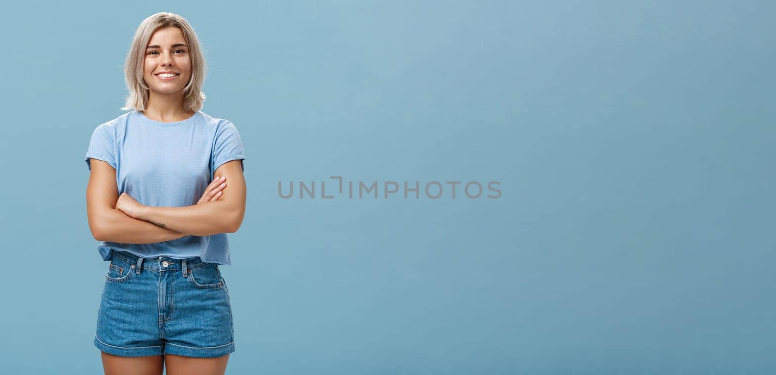 Ambitious good-looking young tanned female with blond hair standing in confident pose with hands crossed on chest smiling broadly standing in denim shorts over blue background. Copy space