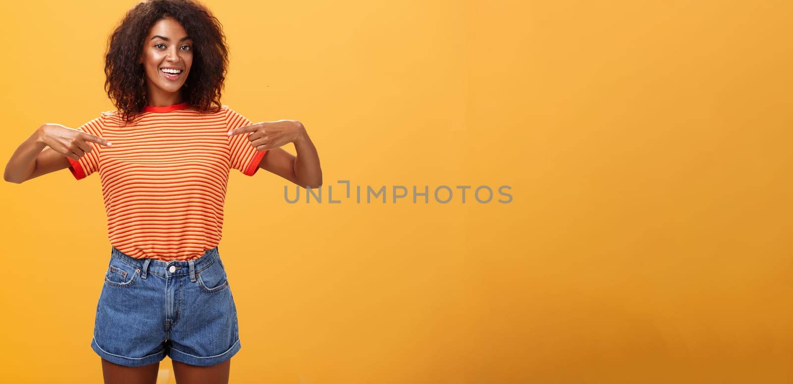 Hey pick me I your girl looking for. Portrait of charming friendly-looking ambitious dark-skinned female with afro hairstyle pointing at chest proudly and joyful posing against orange background.