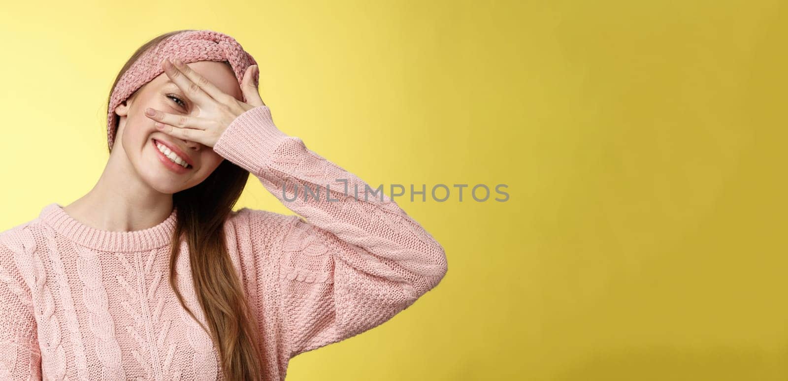 Blushing cute girl feeling positive, smiling happy joyful covering eyes with palm peeking through fingers delighted, playful, tilting head tender and lovely gazing at camera against yellow background.