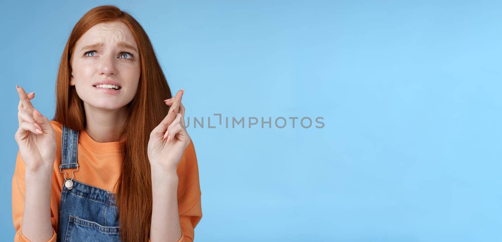 Worried unconfident young redhead intense cute girl frowning nervously waiting improtant moment cross fingers good luck bite lip anxiously frowning look upper left corner praying wish come true by Benzoix