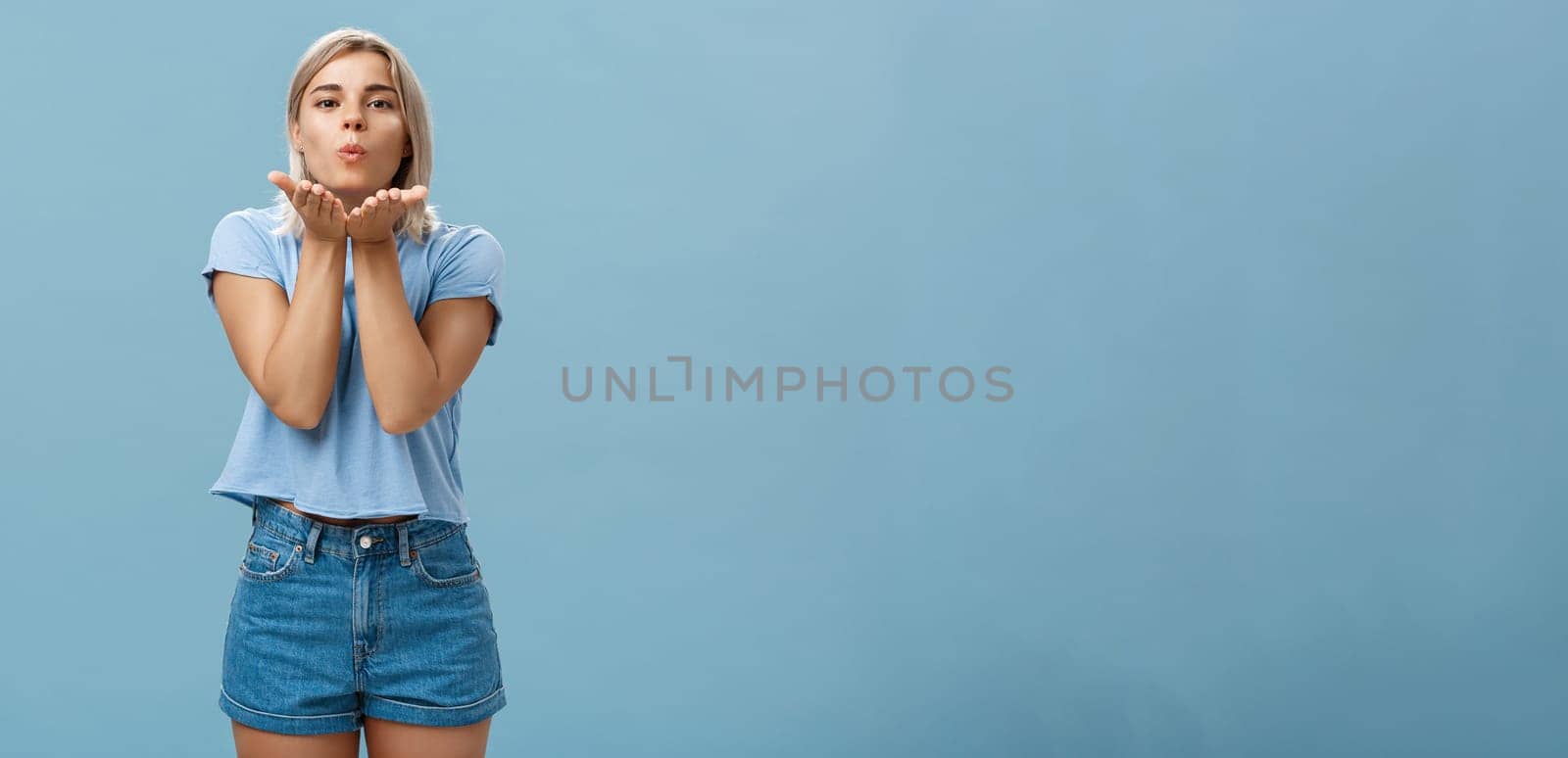 Studio shot of attractive and romantic european blonde girlfriend in denim shorts folding lips sending wind kiss with palms near mouth gazing tender and cute at camera over blue background by Benzoix
