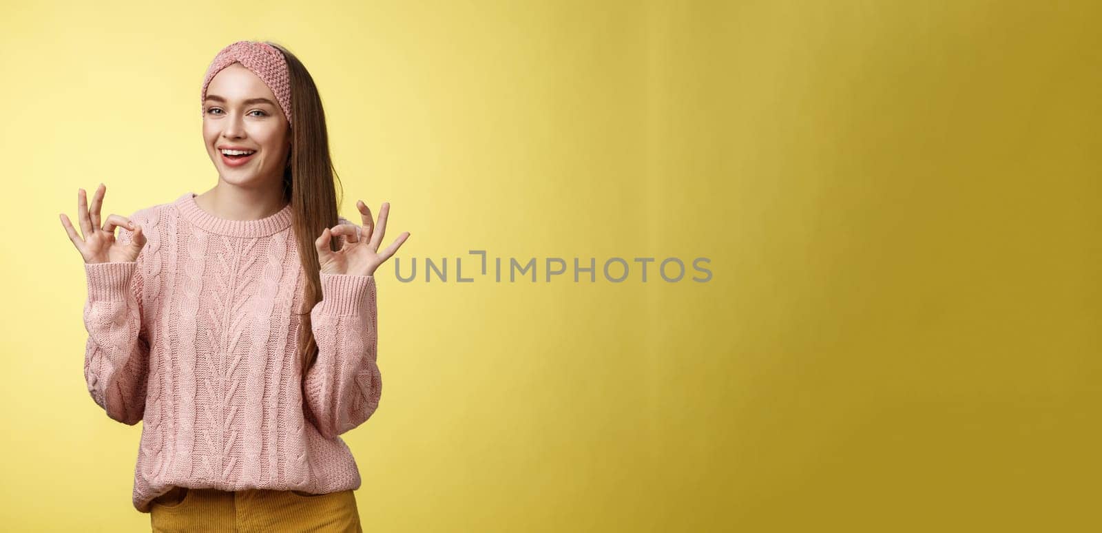 Positive attractive upbeat young cute european woman in 20s wearing casual sweater, headband showing okay, ok gesture smiling assuring everything, good, fine smiling confident over yellow wall.