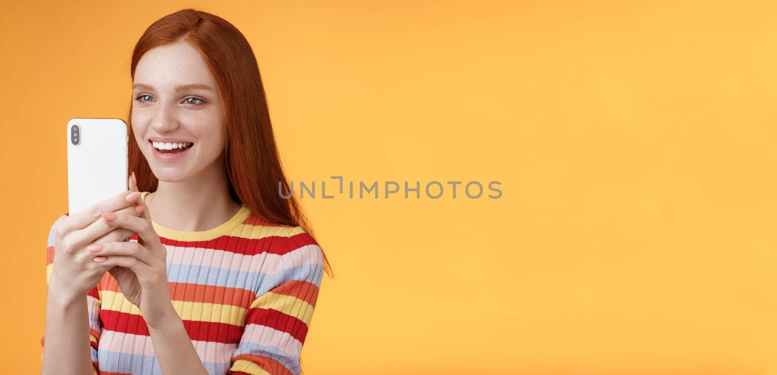 Redhead girl having fun recording hilarious friend actions hold smartphone look display amused shooting funny video telephone standing orange background satisfied smiling delighted. Lifestyle