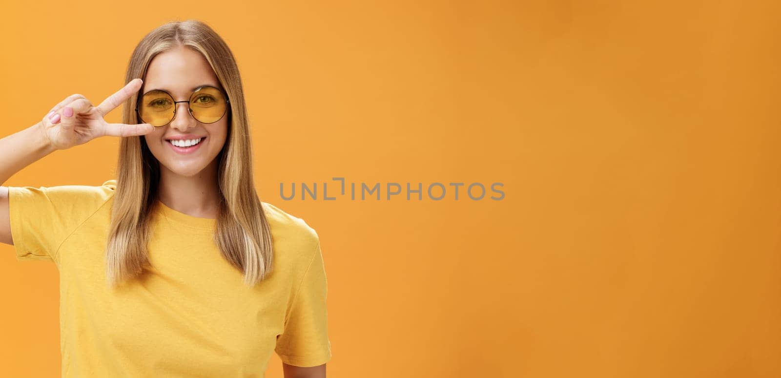 Cute optimistic and friendly young caucasian woman with fair hair in yellow t-shirt and sunglasses showing peace sign near eye and smiling cheerfully at camera taking shot over orange background by Benzoix