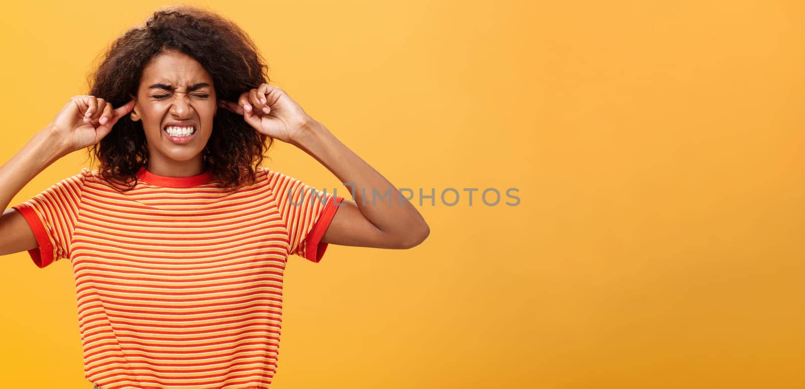 Waist-up shot of intense bothered dark-skinned woman clenchign teeth from discomfort and antipathy closing ears not hear botherind loud noise standing displeased and annoyed over orange wall. Copy space