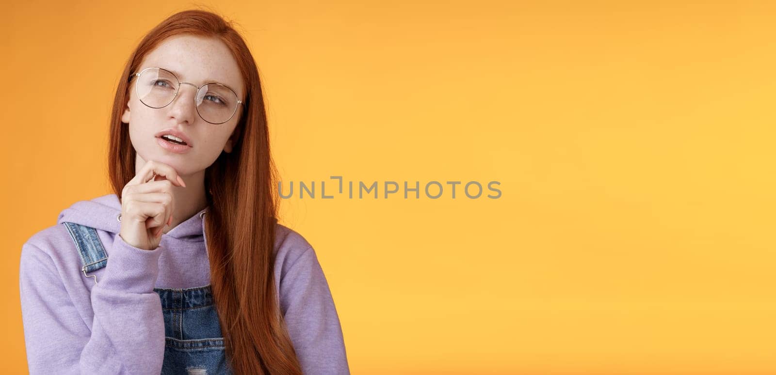 Thoughtful young creative smart redhead girl thinking figure out important thing standing upper left corner squinting thinking get clue touch chin thinking, pondering choice, orange background.