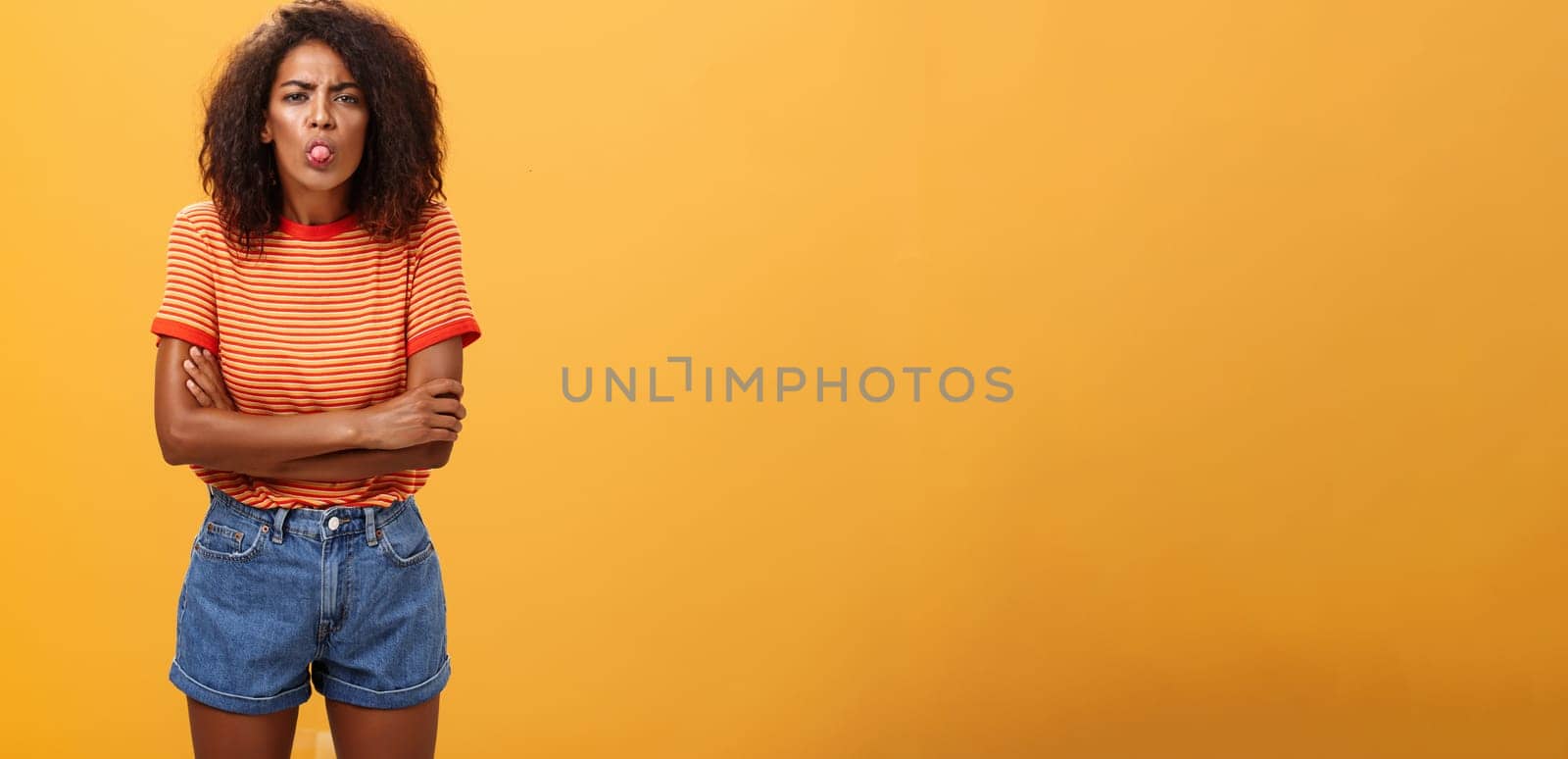 Immature girl showing bad side of character. Portrait of childish offended or displeased young African-American woman with curly hair showing tongue crossing arms on chest over orange background.