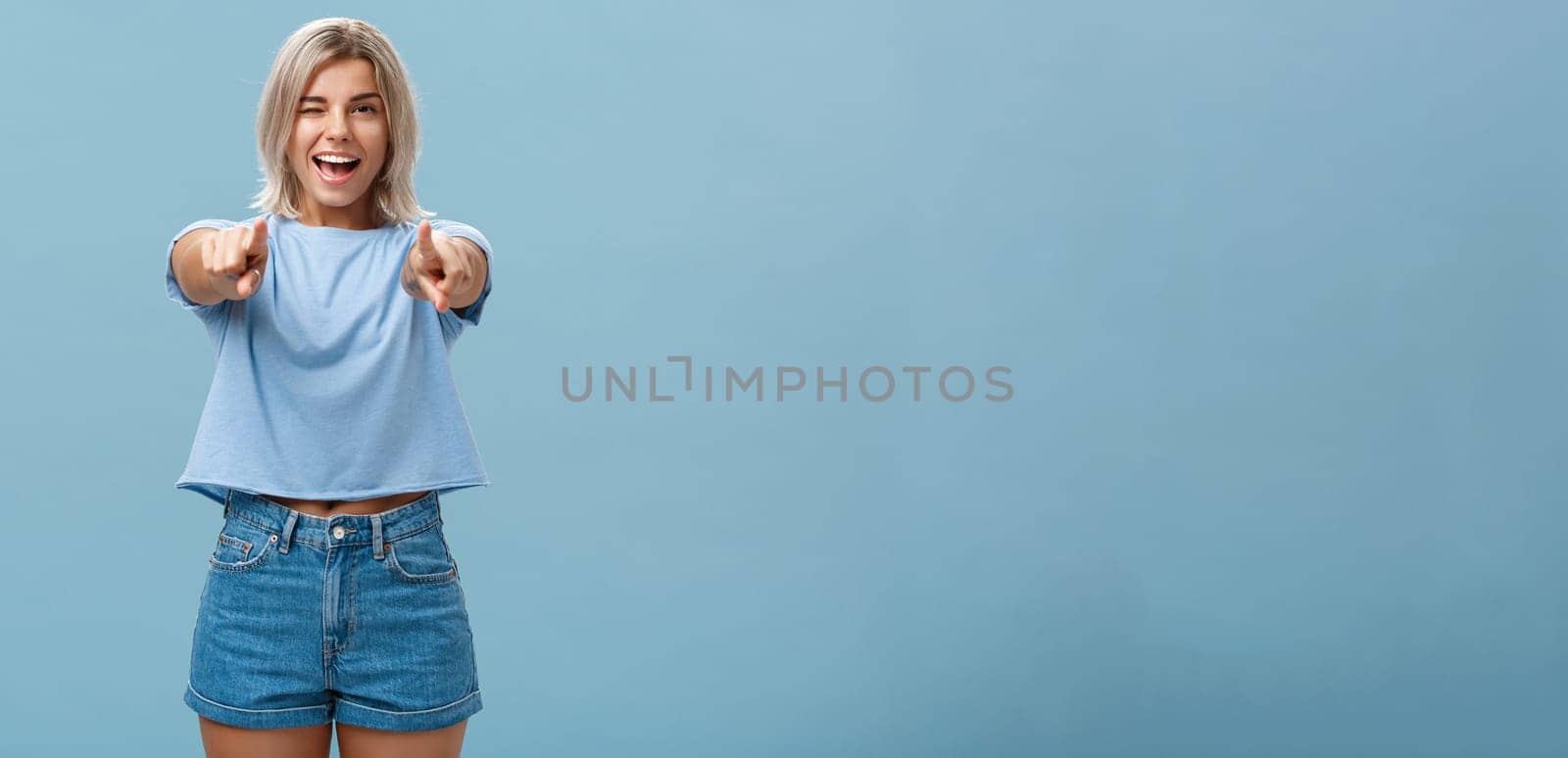 Happy enthusiastic stylish blonde female with tattoos pointing at camera as if picking or making hint smiling joyfully and winking from amazement and happiness posing over blue background by Benzoix