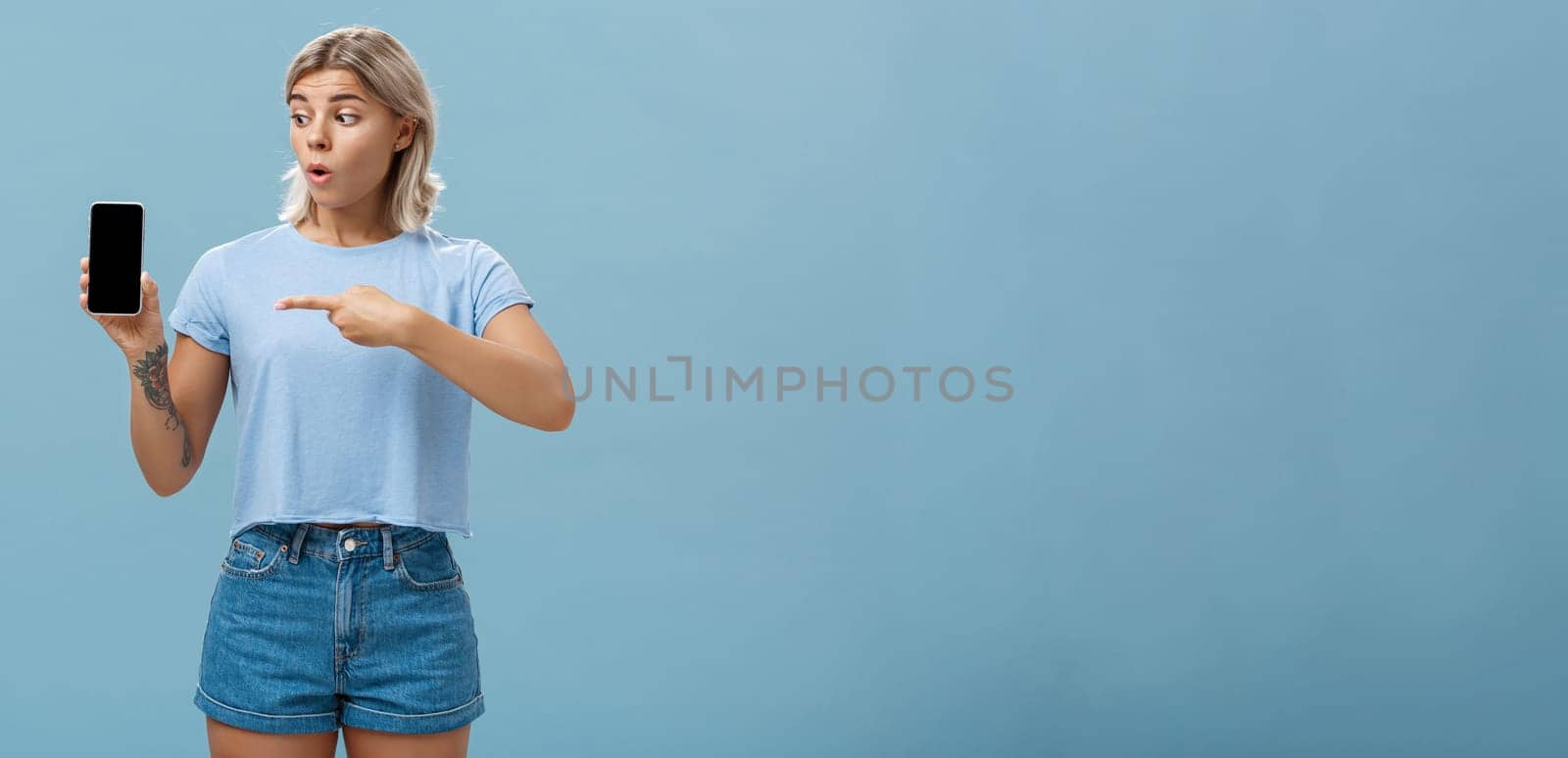 You should download app. Portrait of amazed shocked attractive blonde girl in trendy summer t-shirt holding smartphone gasping from surprise pointing at gadget screen posing over blue background by Benzoix