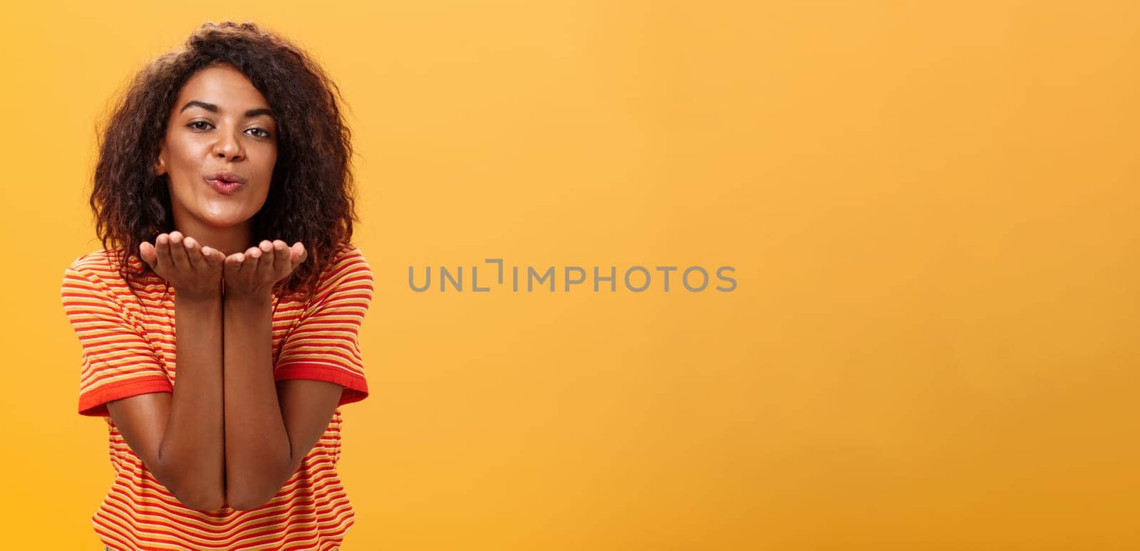 Sending passionate kiss to most loving person. Romantic attractive and stylish young african american girlfriend with curly hairstyle bending towards camera with slight smile, folded lips blowing mwah.