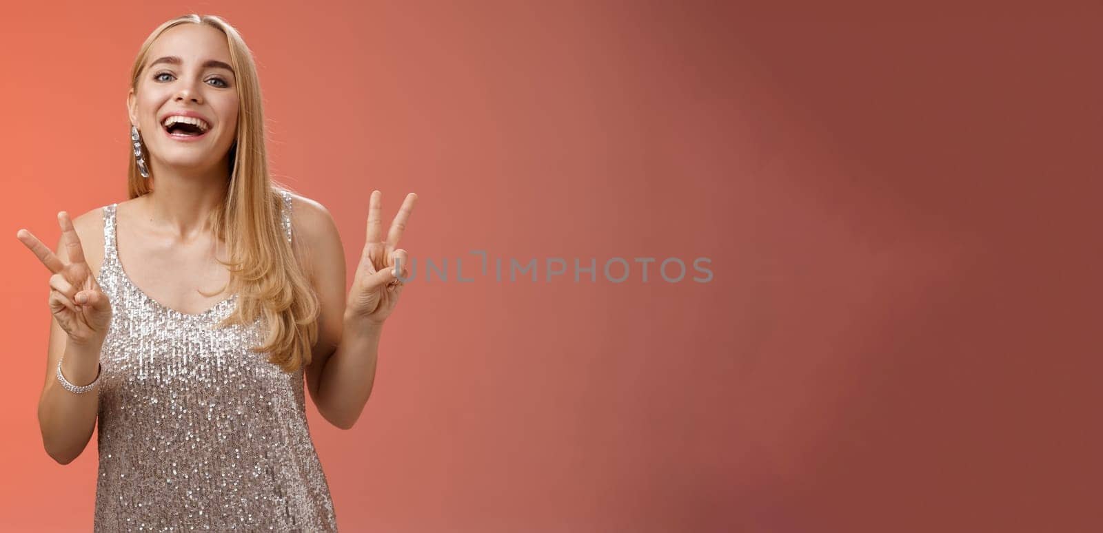 Funny carefree charming caucasian blond female model in silver stylish glamour dress show peace victory gesture smiling broadly have fun celebrating birthday party, standing red background.