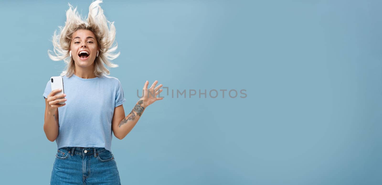 Expressing happiness with help of great tunes. Joyful amused and happy good-looking young female student jumping having fun listenign music in wireless earbuds, holding smartphone over blue wall.