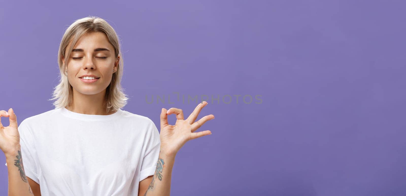 Peaceful calm good-looking female blonde in white t-shirt closing eyes smiling relieved and happy feeling satisfied with life standing in lotus pose with zen gesture over purple background by Benzoix