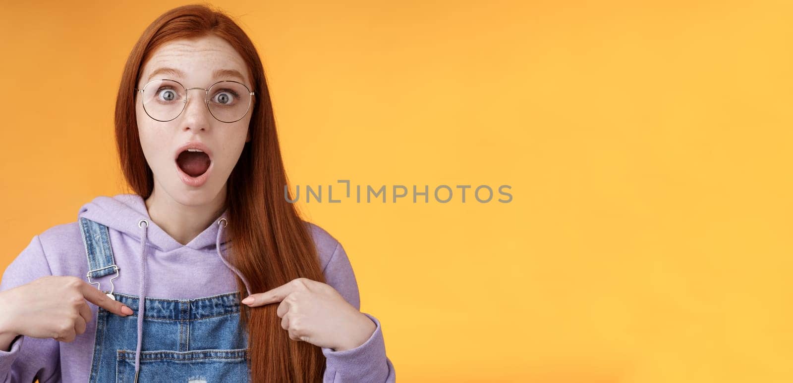 Shocked emotive surprised redhead european woman wearing glasses hoodie drop jaw gasping stunned pointing herself full disbelief cannot believe win being picked look impressed orange background by Benzoix