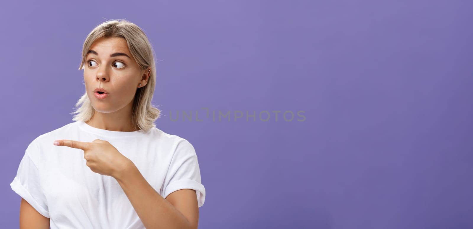 Close-up shot of intrigued interested attractive adult blonde female in white t-shirt folding lips staring and pointing left with curious and thrilled expression over purple background by Benzoix