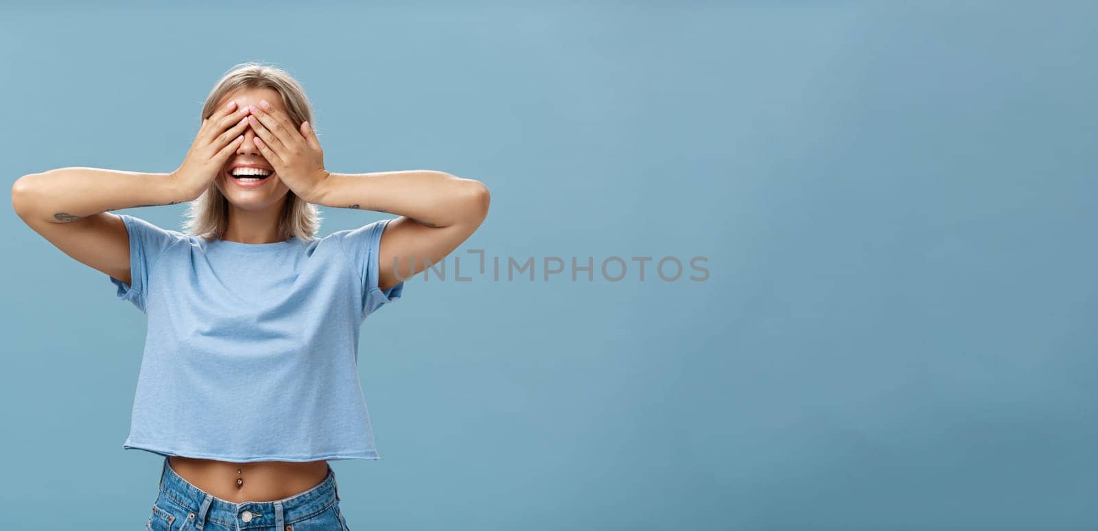 Hide if you can. Portrait of carefree and happy good-looking playful girl with blonde hair smiling broadly while counting ten and playing hide-and-seek with closed eyes over palms near blue wall.