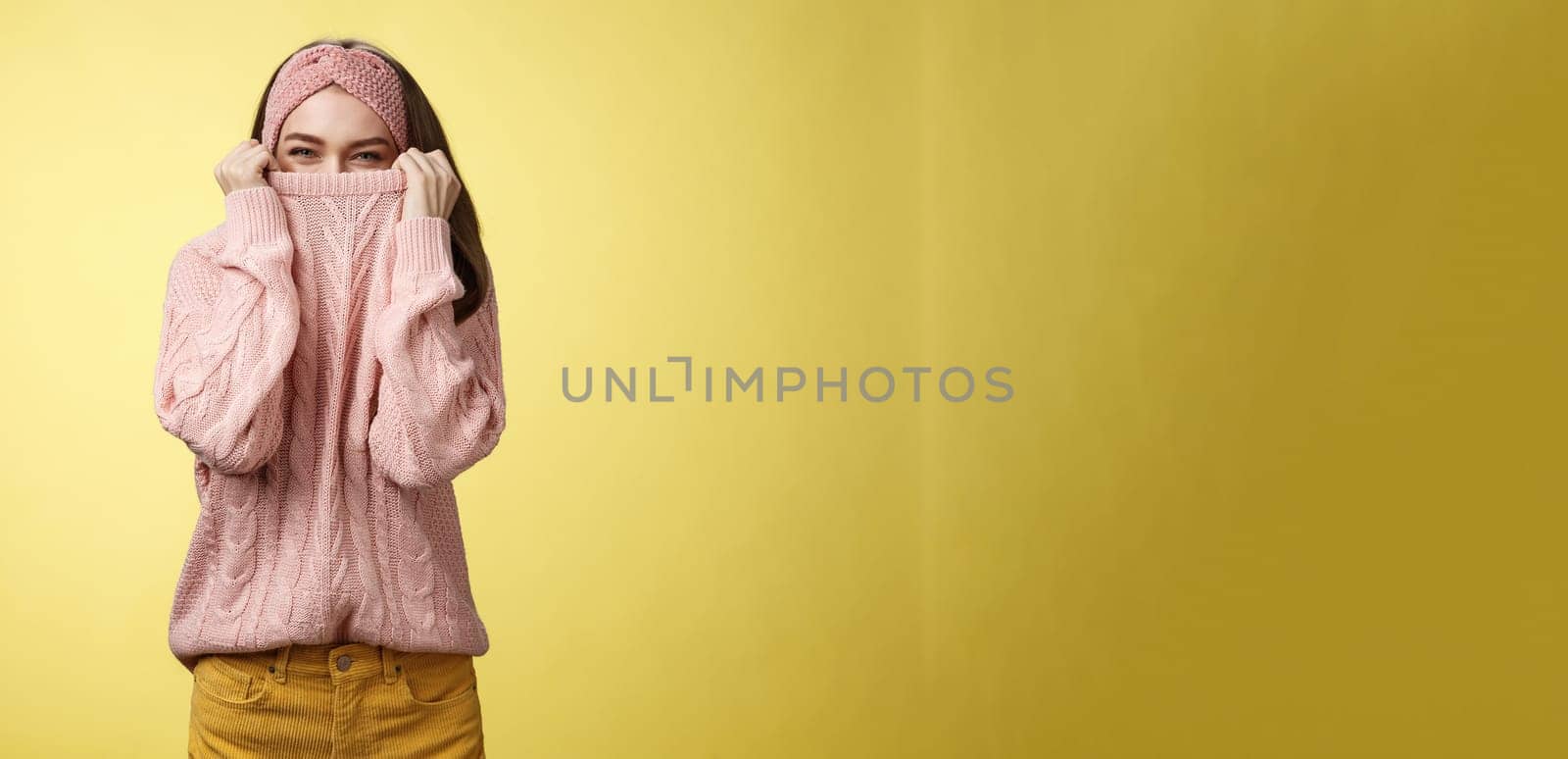 Cute playful young girl hiding in sweater peeking and squinting flirty over top of cloth pulling collar on nose, fooling around having fun happily, being in positive good mood, warming-up on cold day.