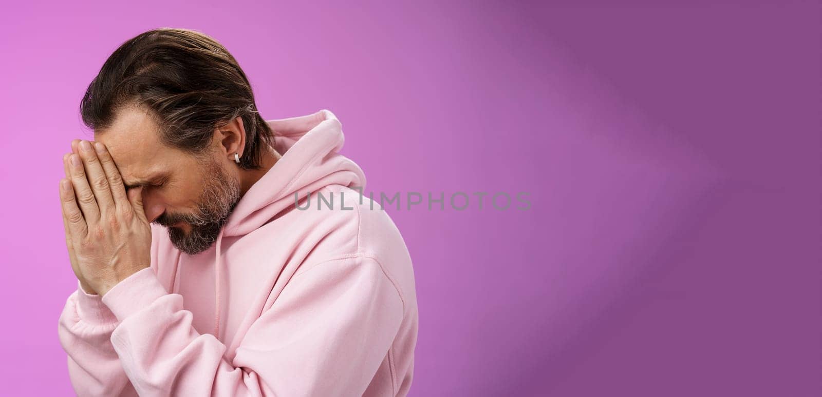 Concerned upset man supplicating asking god mercy help praying bow head close eyes hold hands pray hopefully waiting miracle worried wife health, standing sadness purple background. Copy space