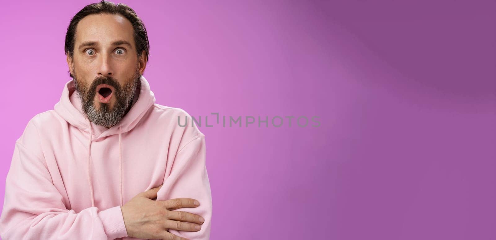 Shocked impressed gossiping caucasian bearded adult 40s man grey hair in pink hoodie gasping fascinated fold lips wow widen eyes astonished hearing interesting thrilling story, purple background.