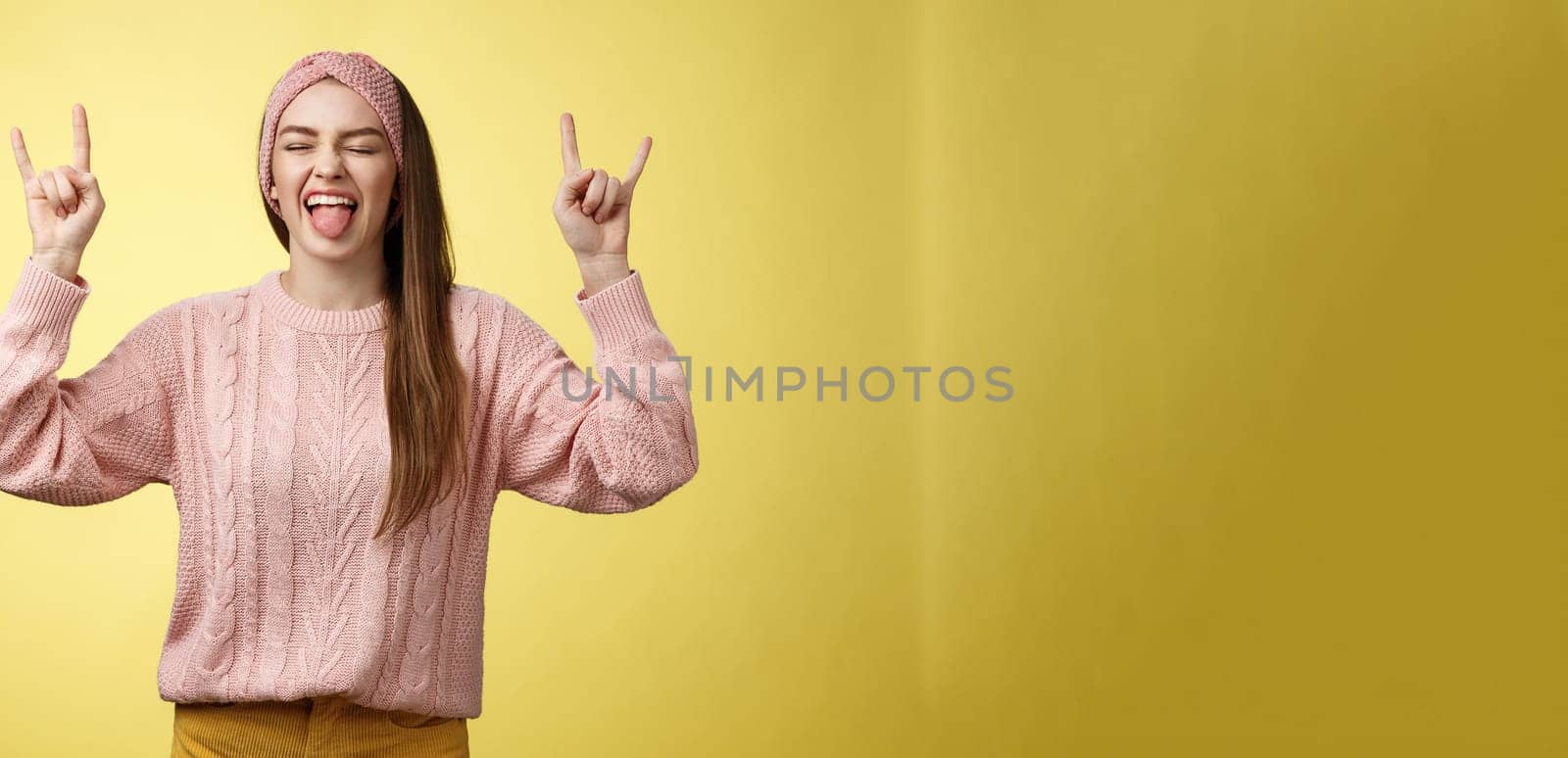 Lifestyle. Cute heavy metal lover showing rock roll symbol sticking tongue amused and happy fooling around listening favourite music posing excited and pleased against yellow background in knitted outfit.