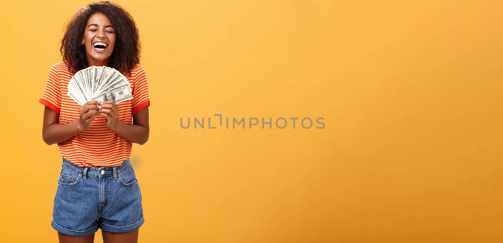 Finally girl rich. Portrait of happy charismatic stylish young female model receiving first payment holding lots of money and laughing from joy winning lottery ready waste cash over orange background.