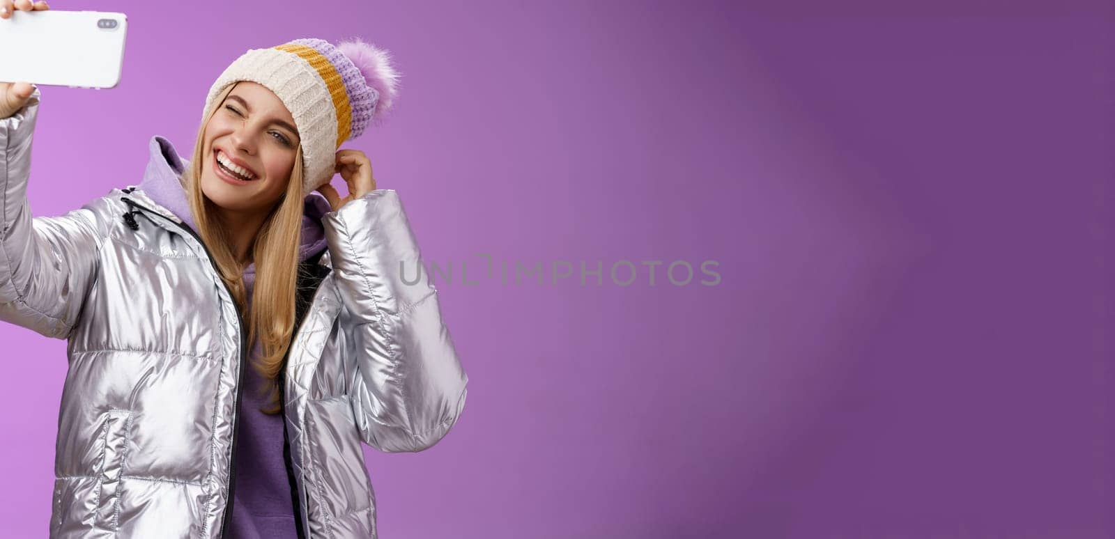 Joyful carefree charming blond girl having fun wanna post pictures from ski resort vacation taking seflie holding smartphone mimicking show tongue winking look mobile display, purple background by Benzoix