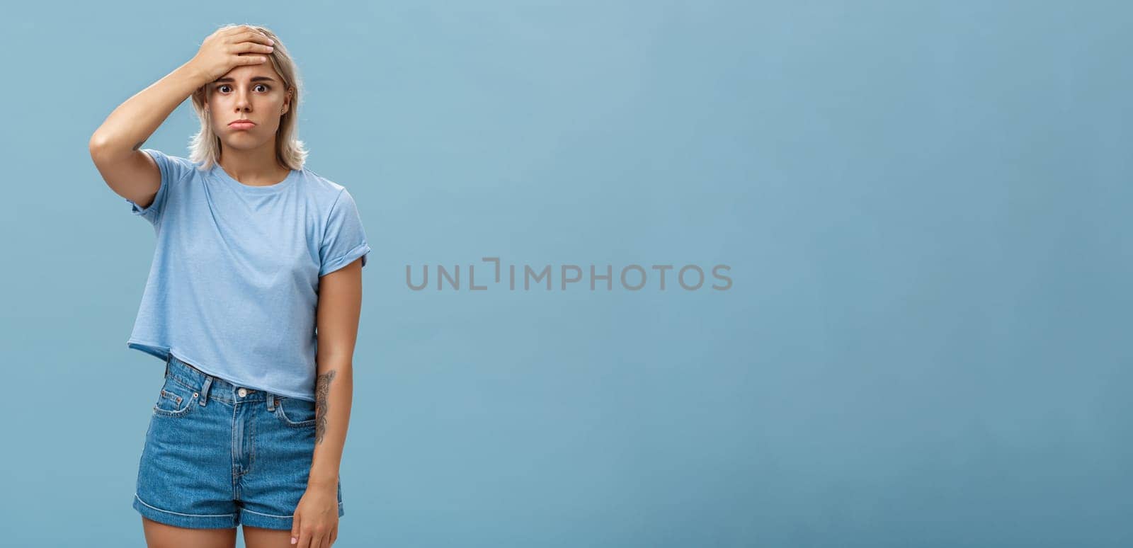 Concerned and troubled young teenage girl being shocked with loads of responsibilities holding hand on forehead staring spaced out and pouting from tiresome perplexed situation over blue wall by Benzoix