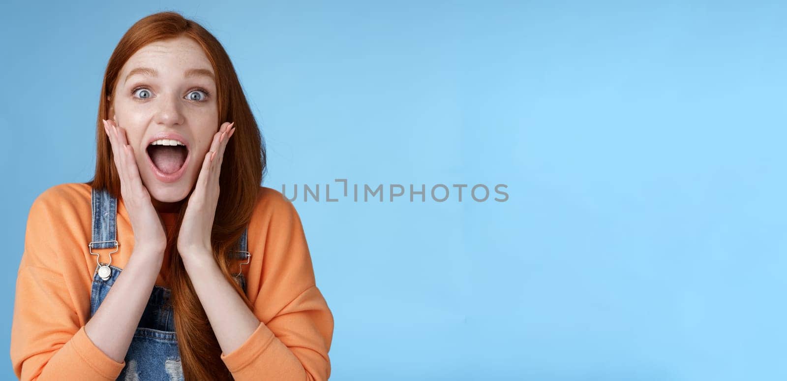 Excited thrilled young emotional enthusiasitc ginger girl teenage college student yelling amused smiling broadly receive positive good news look surprised camera touch face astonished blue background by Benzoix