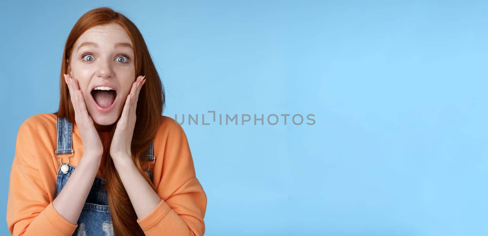 Lifestyle. Excited thrilled young emotional enthusiasitc ginger girl teenage college student yelling amused smiling broadly receive positive good news look surprised camera touch face astonished blue background.