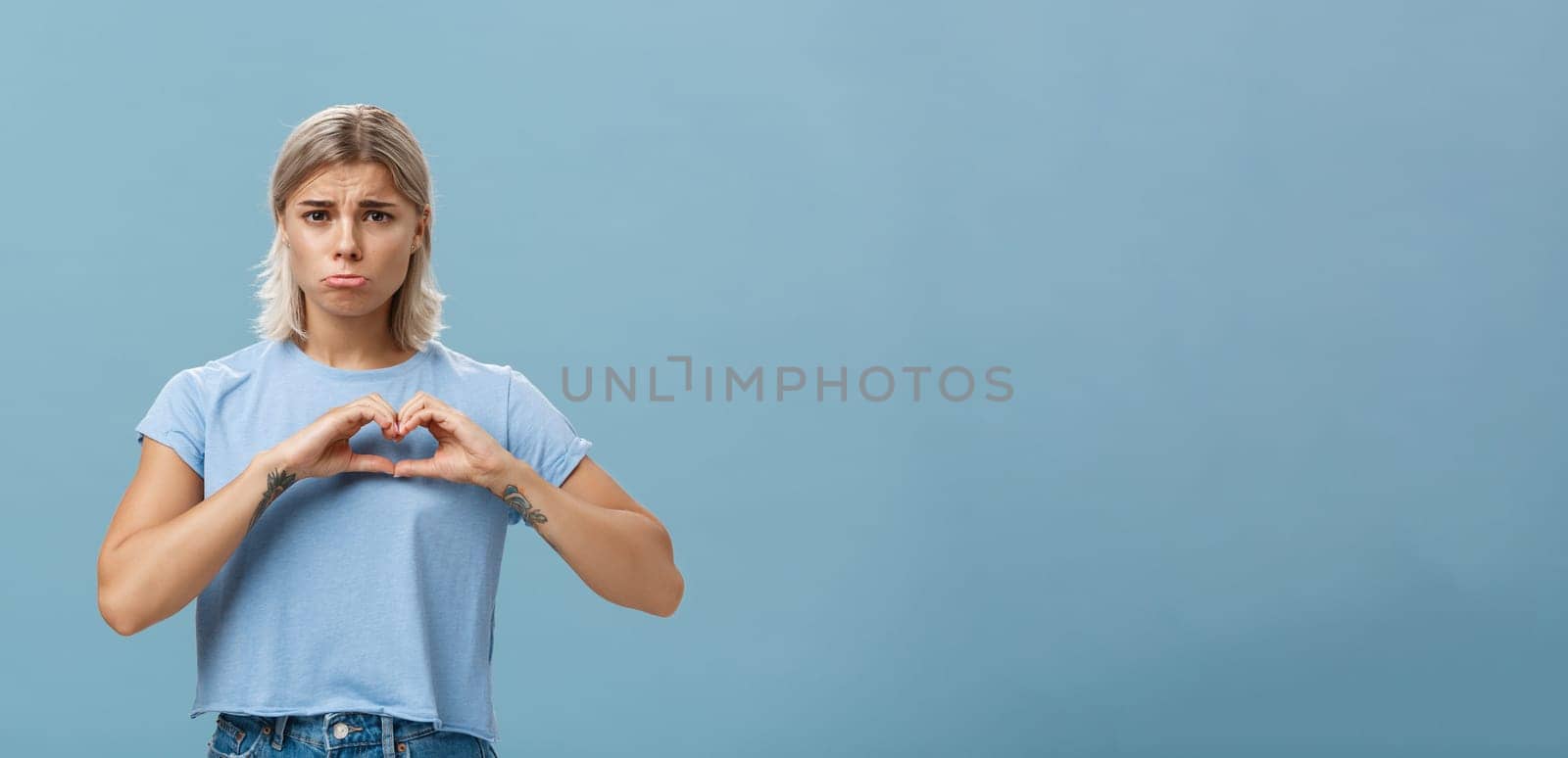 Heart being broken. Sad and gloomy heartbroken girl with blond hair tattoos on arms and tanned skin pursing lips whining and complaining making love sign over breast standing unhappy near blue wall.