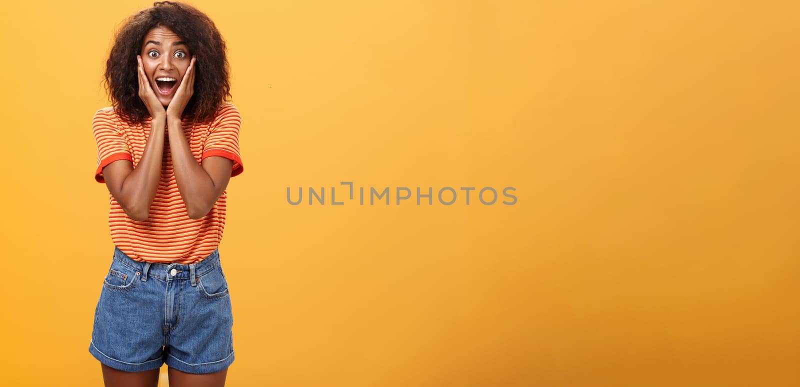 Impressed talkative african american woman finding out hot details of rumor being excited, thrilled leaning face on palms, smiling broadly with amused expression listening carefully over orange wall.