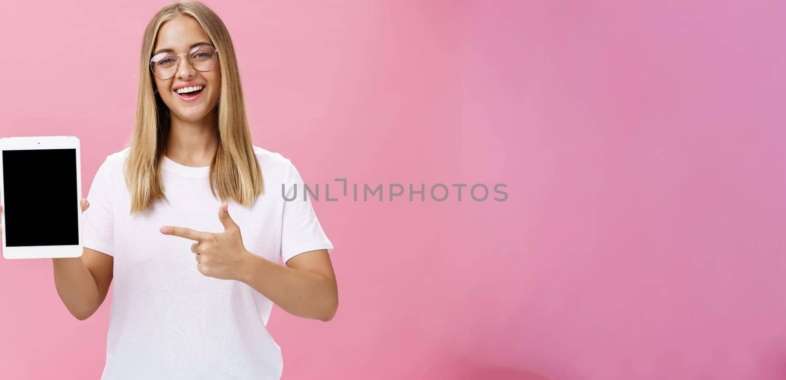 Female freelance programmer proudly showing her app for digital tablet holding gadget pointing at device screen smiling broadly with delighted expression wearing glasses, posing over pink wall by Benzoix