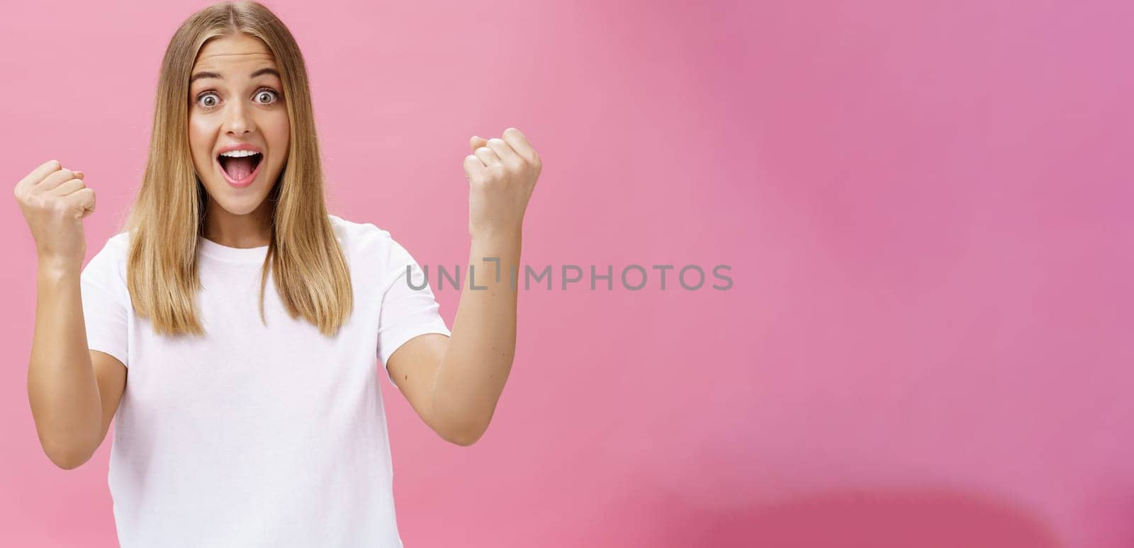 Excited cheerful and optimistic charming woman with fair hair in white t-shirt raising fists in victory and triumph yelling yes in success and amazement standing supportive against pink background. Lifestyle.