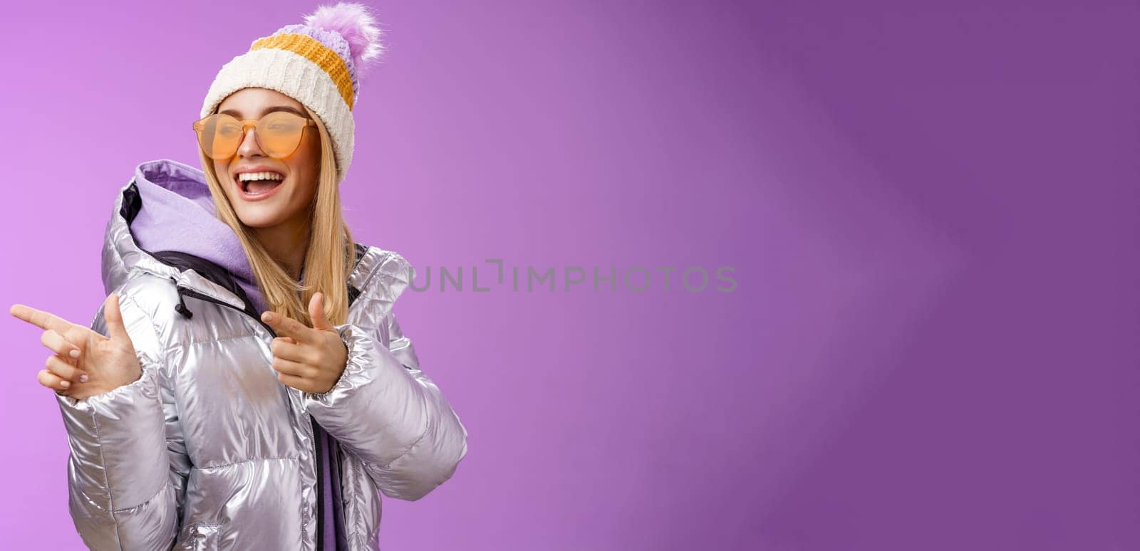 Hey how you doin. Cheeky stylish bond girl having fun greeting friend pointing finger pistols left smiling sassy say hello what up wearing cool silver jacket hat sunglasses, purple background.