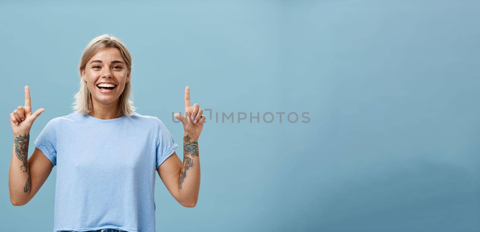 Waist-up shot of upbeat creative stylish and gorgeous blonde female student have tattoos on arms pointing up with raised forefingers smiling with delight and joy posing over blue background. Advertisement concept