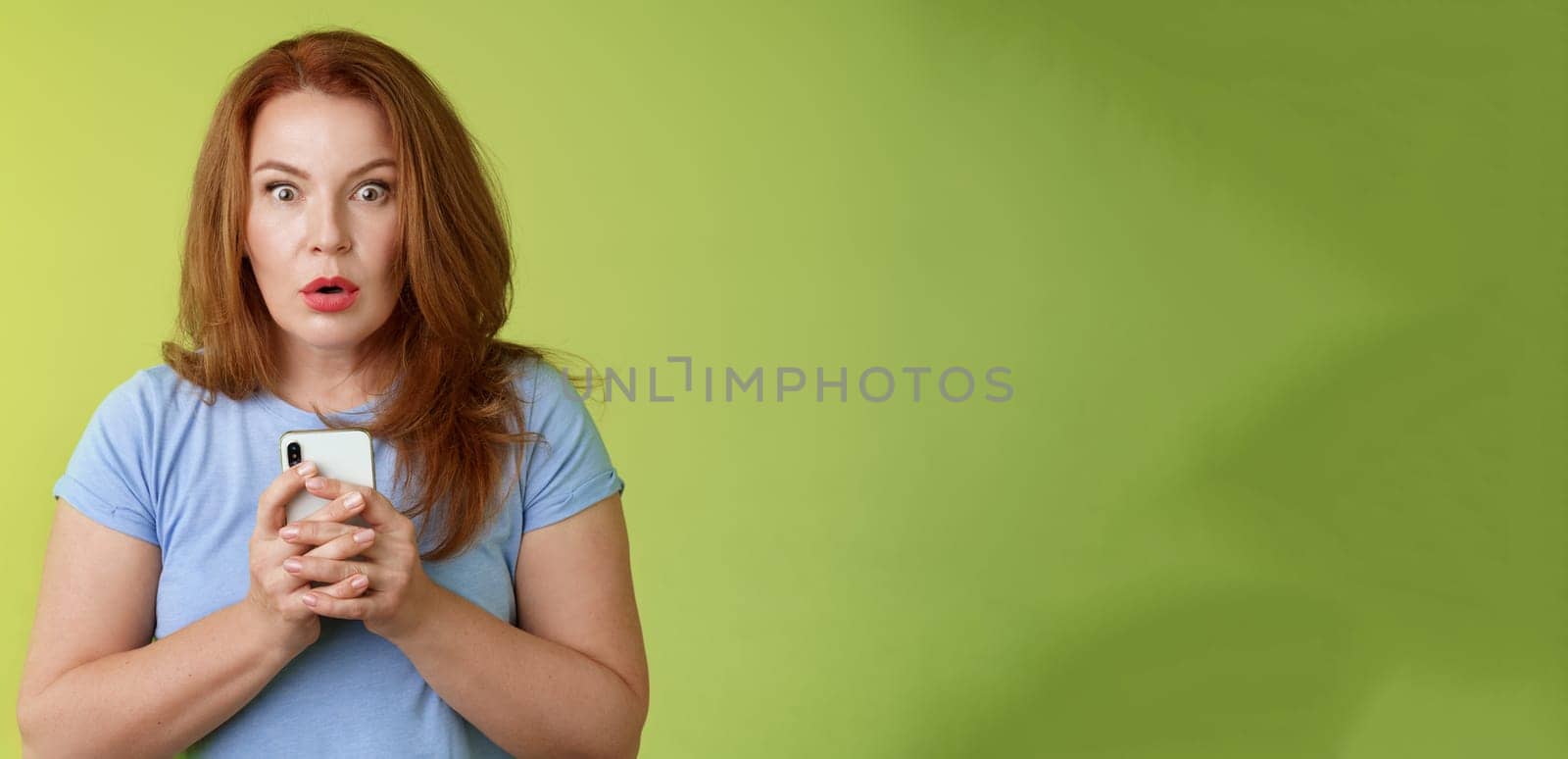 Shocked speechless impressed redhead european middle-aged woman fold lips astonished wow stare camera fascinated react amazed wondered received message hold smartphone intense green background.