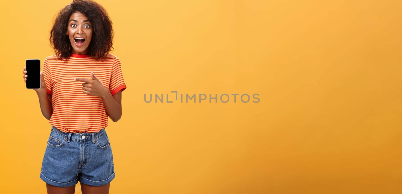 Impressed fascinated stylish slim african american curly-haired woman in striped t-shirt dropping jaw from amazement holding awesome smartphone pointing at device screen over orange wall. Lifestyle.