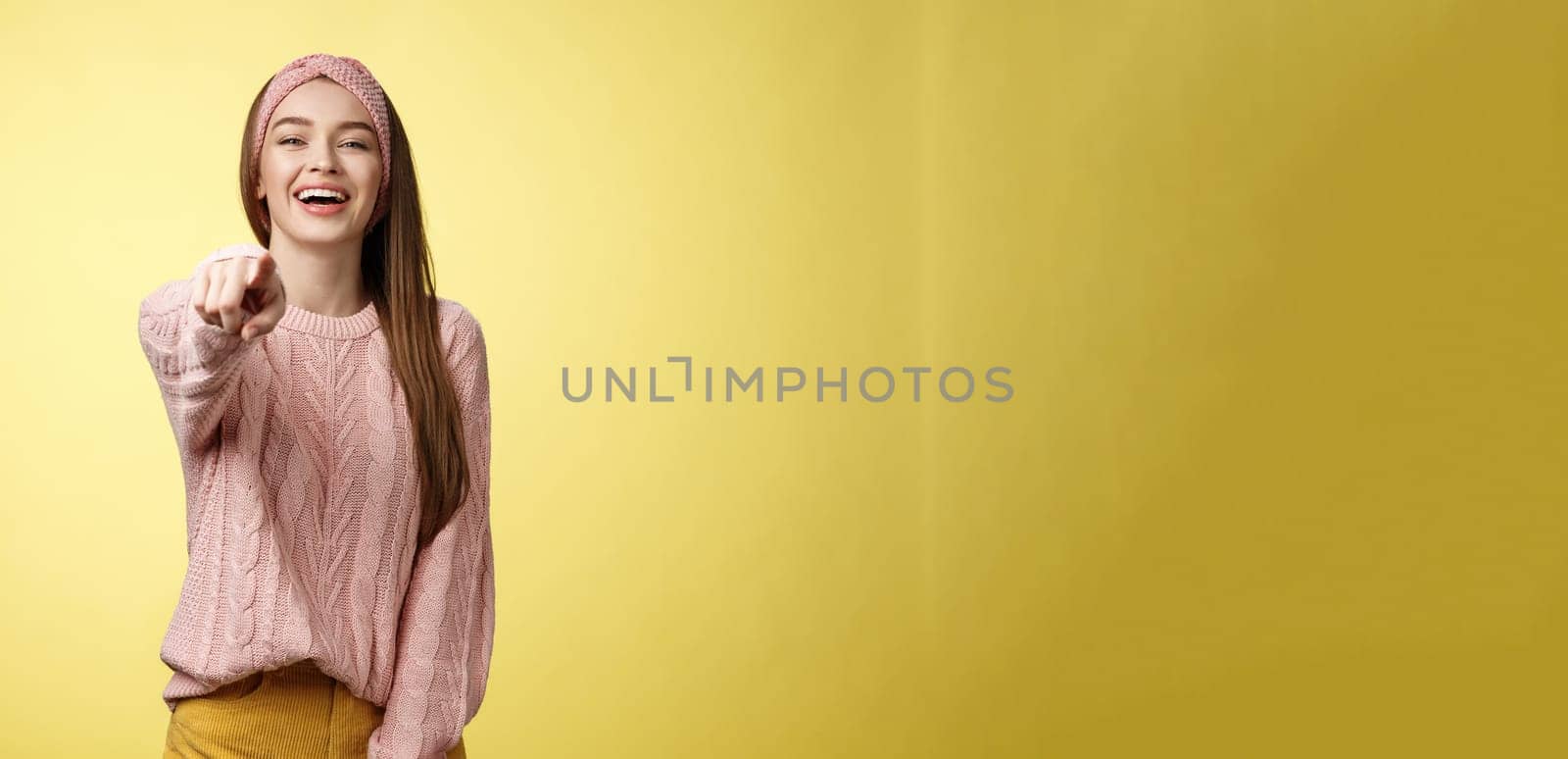 Amused charming tender young european girlfriend mocking having fun of someone pointing finger at camera laughing over funny situation, chuckling, mockering against yellow background. Copy space