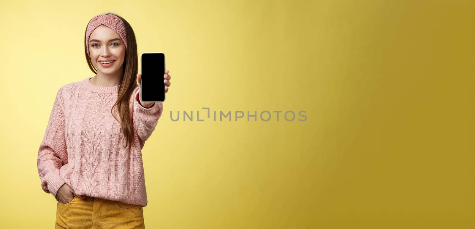 Best smartphone here you go. Charming outgoing young pretty woman in knitted sweater, headband extending hand with phone showing gadget screen smiling recommending cellphone over yellow wall.