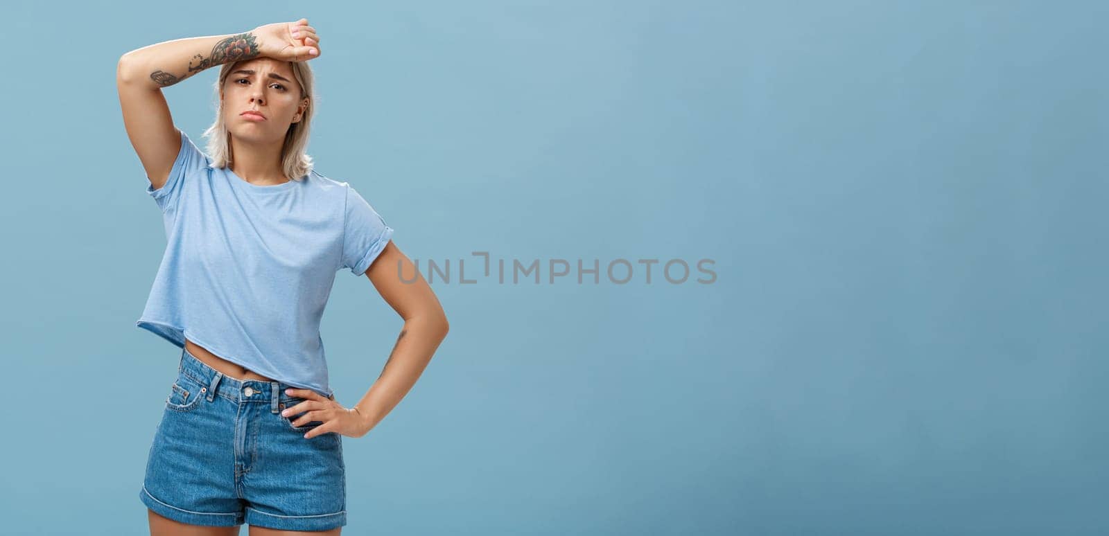 Tired need help save me from work. Gloomy displeased exhausted young sportswoman with tattoos and tanned skin whiping sweat from forehead gazing upset at camera being fed up of problems.