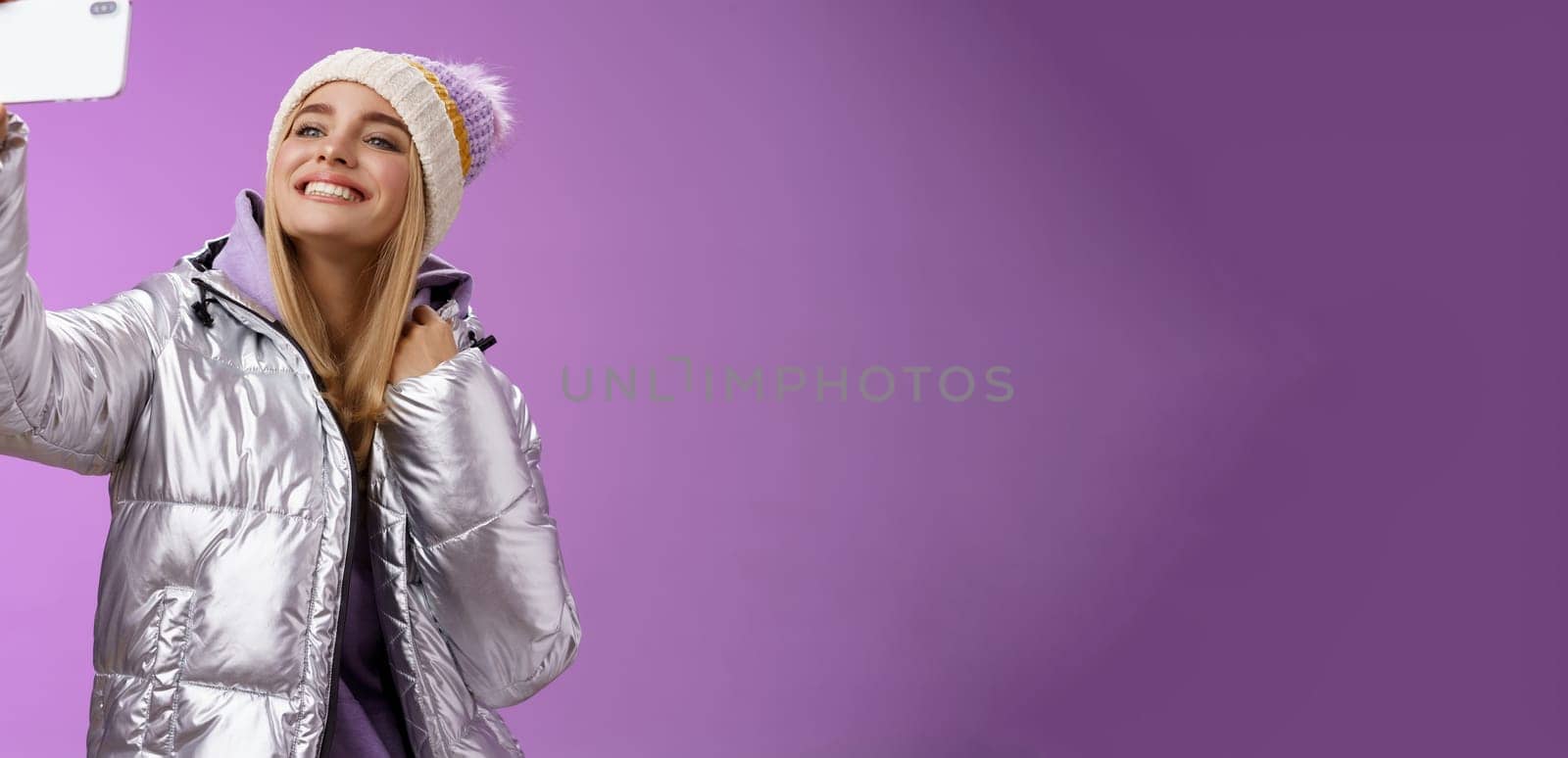 Happy delighted carefree fair-haired charming european woman in silver winter jacket hat raising smartphone horizontally taking selfie smiling mobile phone display, purple background.