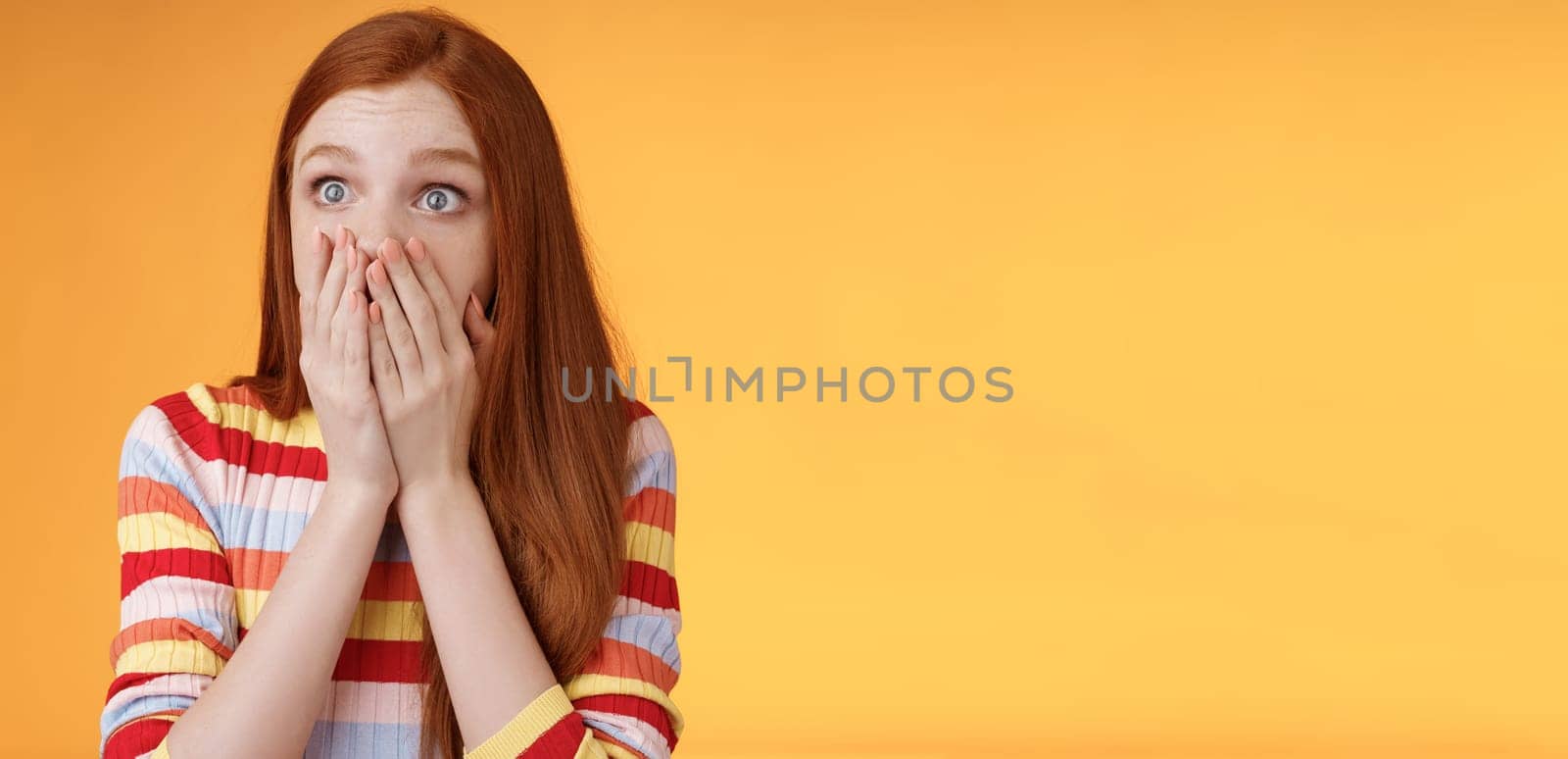 Shocked upset redhead female witness terrible accident gasping cover mouth palms astonished staring left frightened express empathy worry standing orange background panicking. Copy space