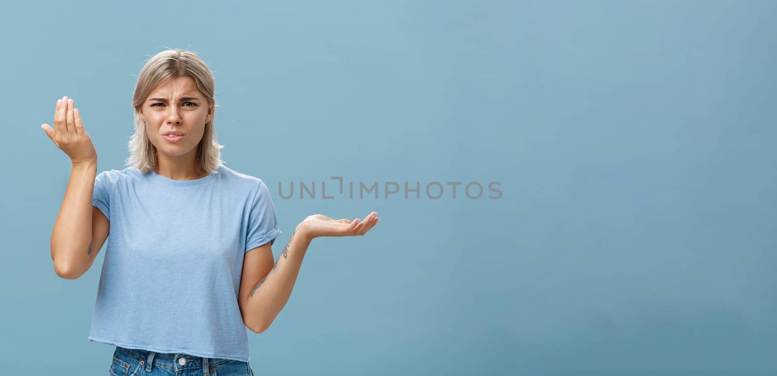 Wtf you saying. Portrait of pissed and confused stylish girlfriend with blond hair raising palm high and aside with perplexed expression being questioned hearing stupid nonsense over blue wall.