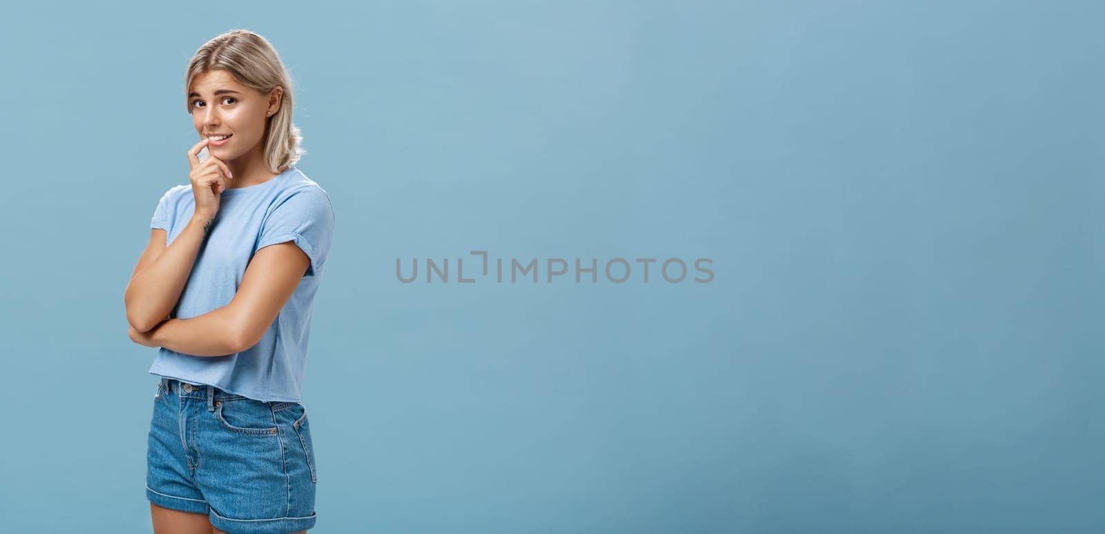 Studio shot of unconfident silly and insecure cute feminine girl with blond hair standing half-turned over blue background holding finger on lip while being shy ask question smiling hesitating by Benzoix