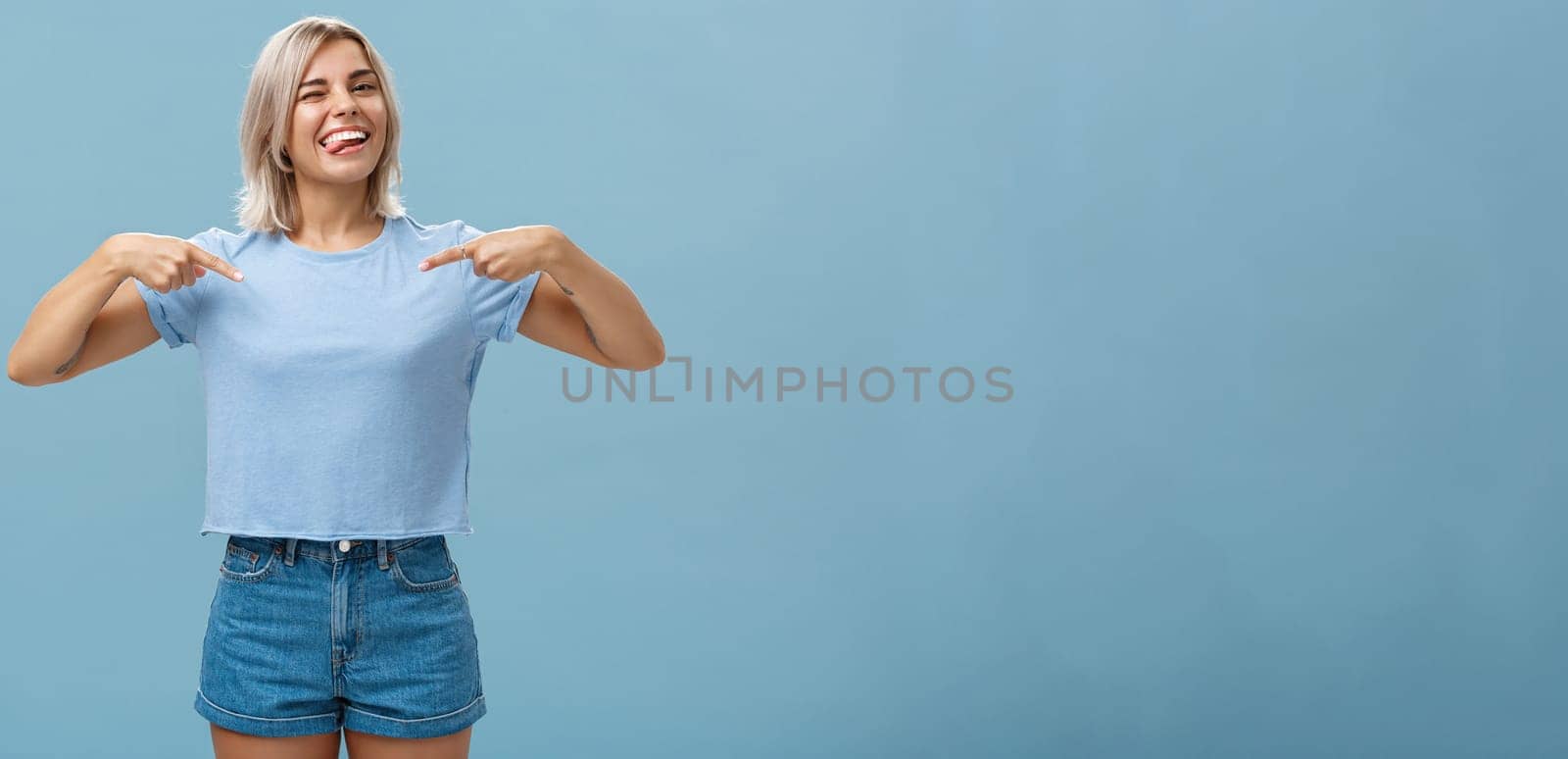 Yeah proud of myself. Self-assured joyful and carefree attractive blonde female with tanned skin and tattoos pointing at herself winking and sticking out tongue with broad smile over blue wall by Benzoix