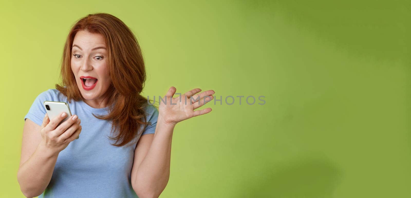 Surprised glad middle-aged pretty redhead woman look smartphone raise hand celebratory excitement open mouth reacting impressed gladly read good excellent news look mobile screen green background by Benzoix